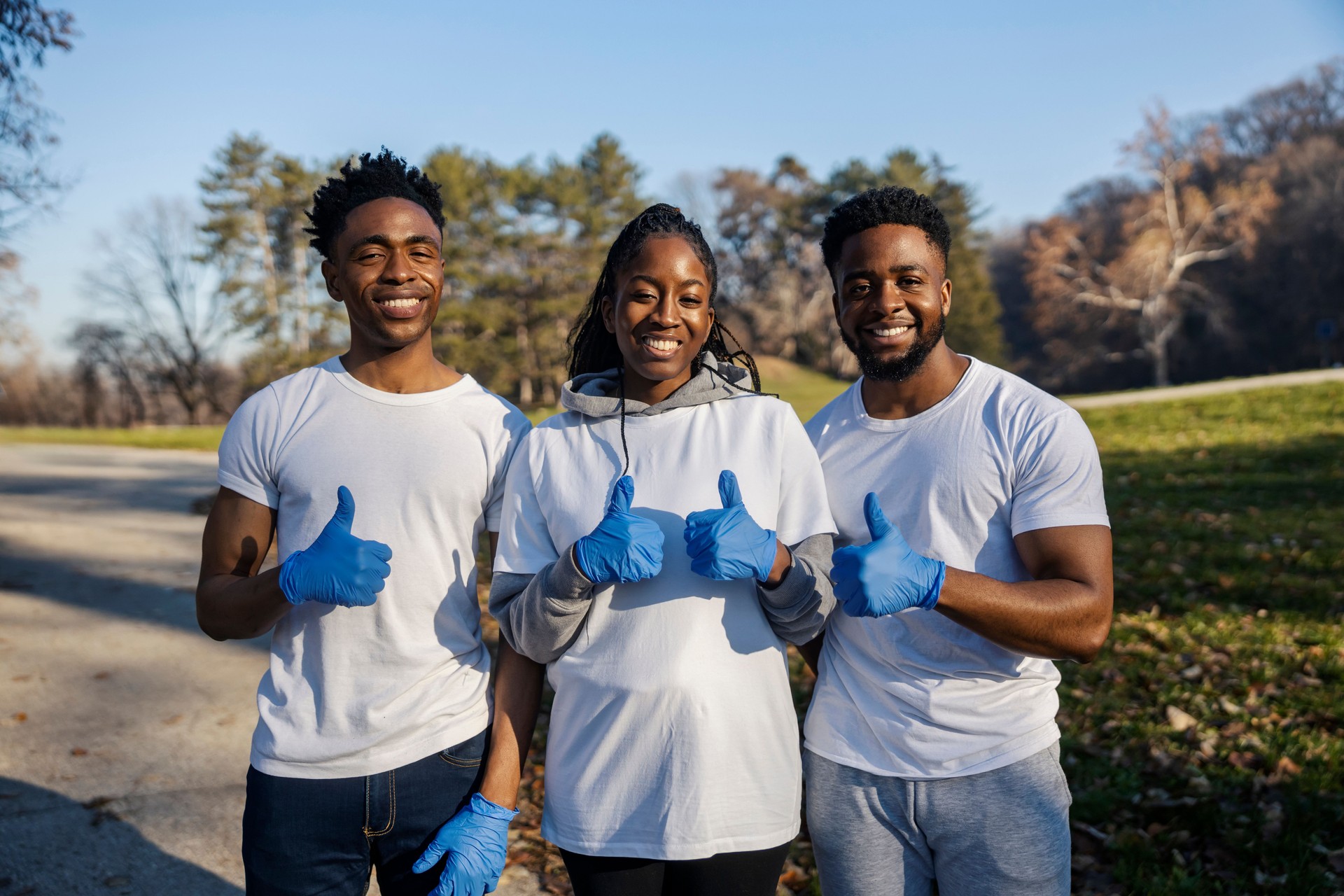 Portrait of three multicultural volunteers standing in nature and giving thumbs up at camera on earth's day.
